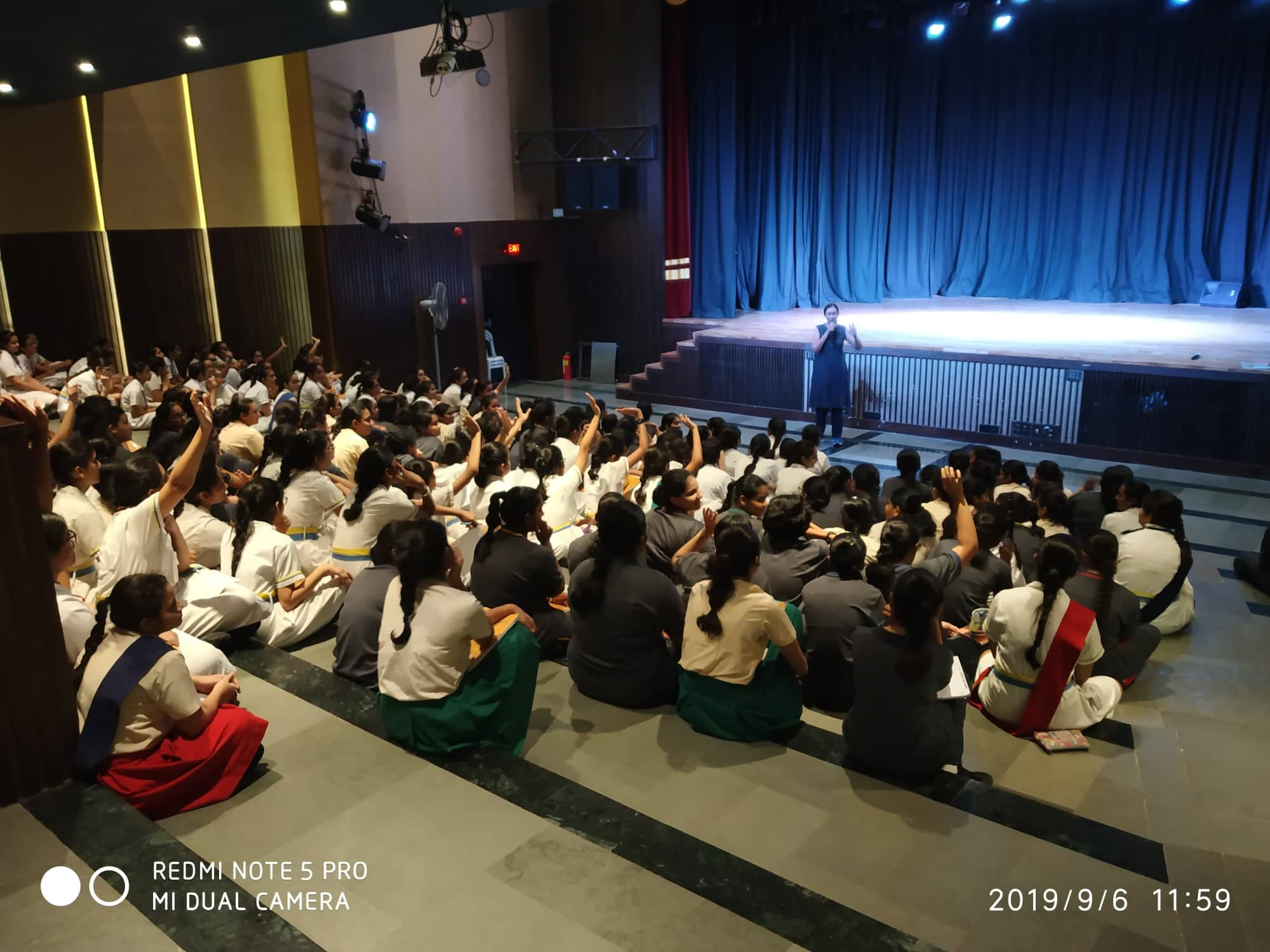 Varsha Seshan speaking on the mic to an audience of schoolgirls, many of who are raising their hands