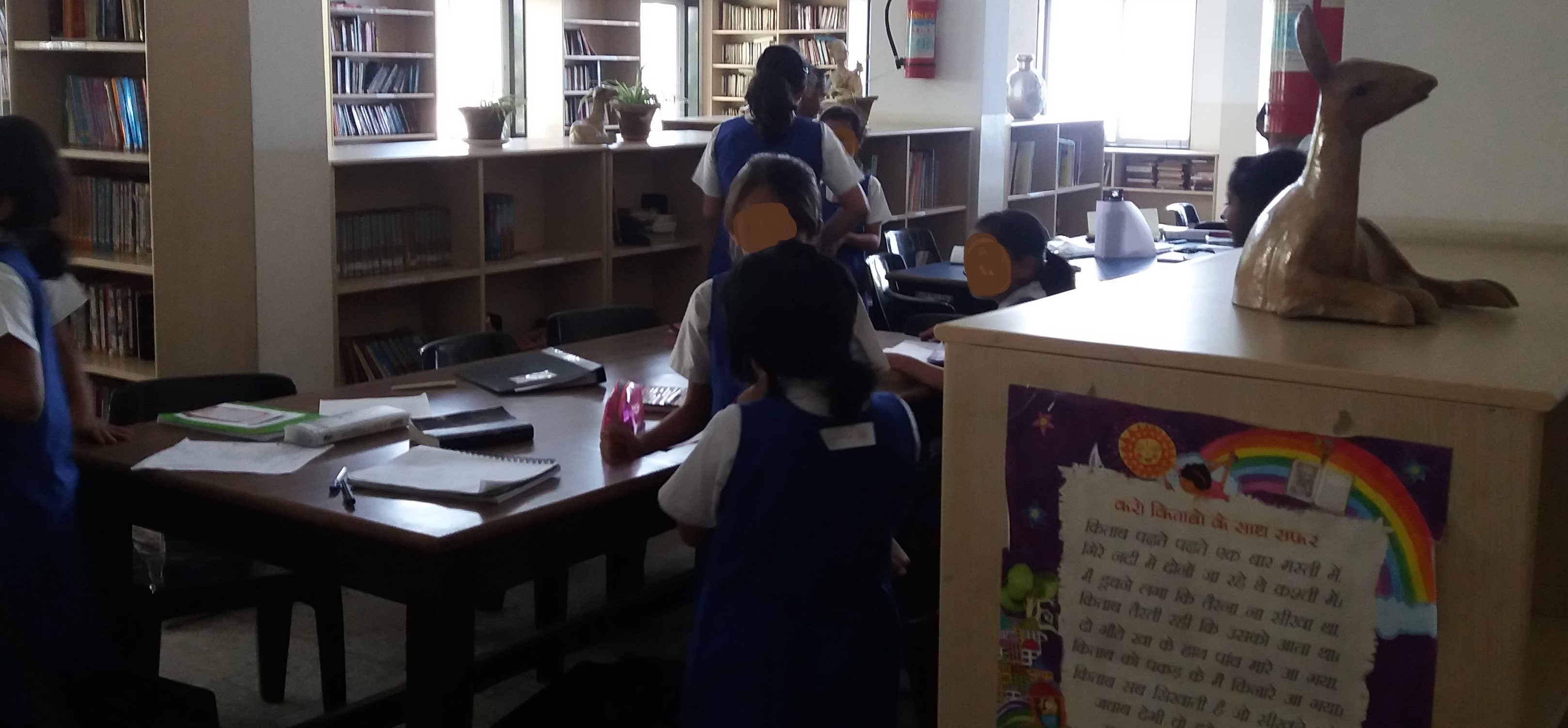 A few girls roaming around in the library, notes pinned to their backs. Faces blurred.