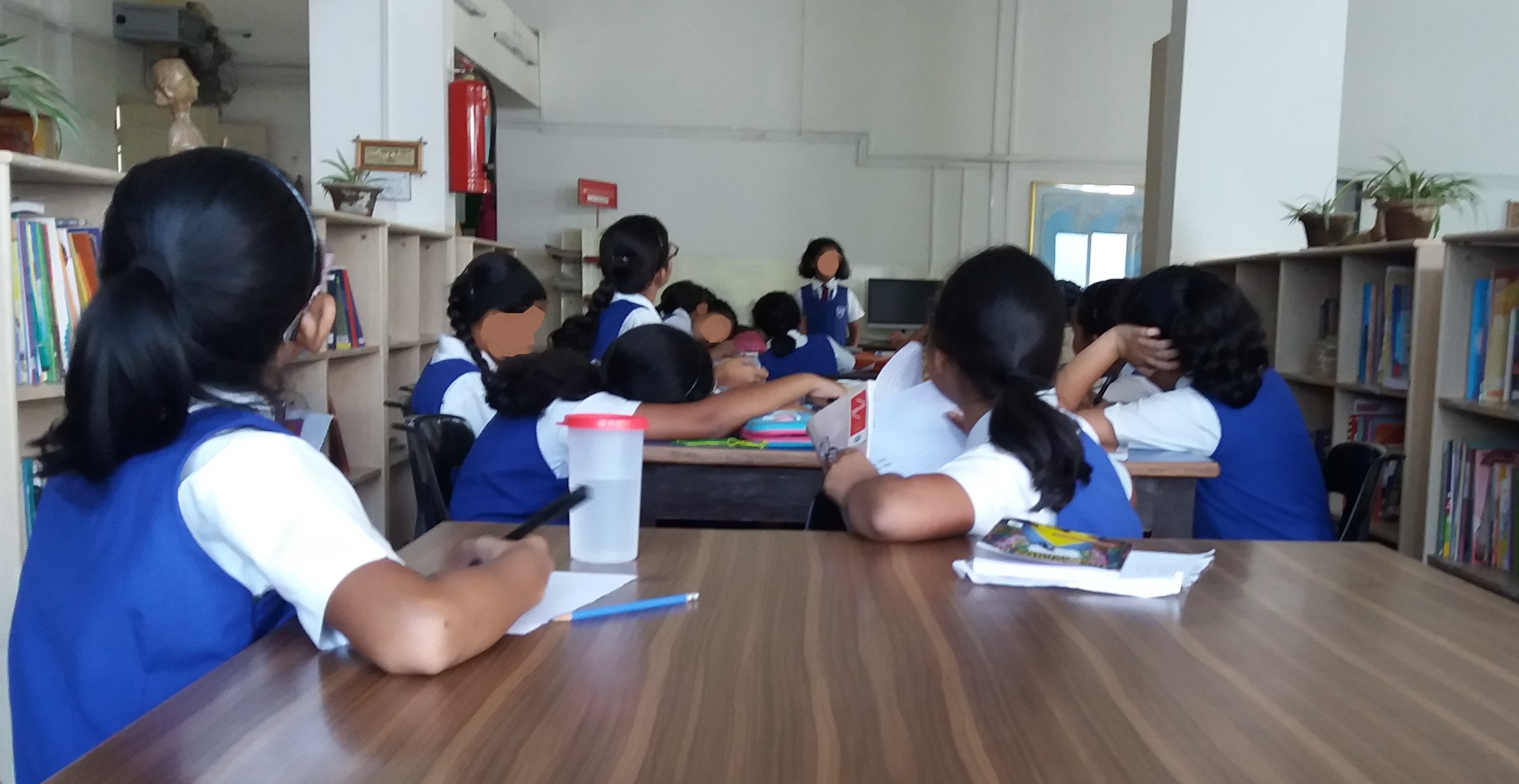 Girls sitting in a library looking ahead at one girl who is standing and speaking. Everyone is in uniform. Faces deliberately blurred.