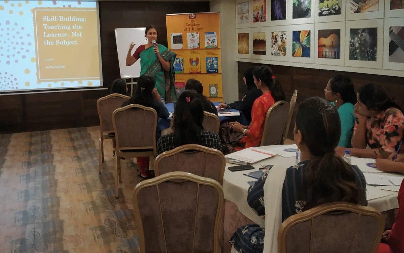 Varsha Seshan speaking on the mic to a group of teachers. In the background, a presentation that says 'Skill-Building: Teaching the Learner, Not the Subject'