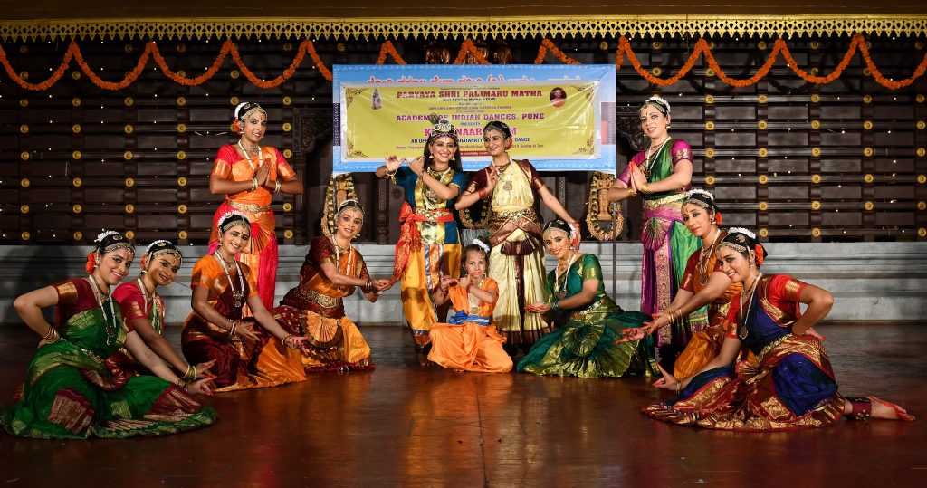 Dancers in Bharatanatyam costume in a semi-circle. In the centre, one dancer dressed as Krishna