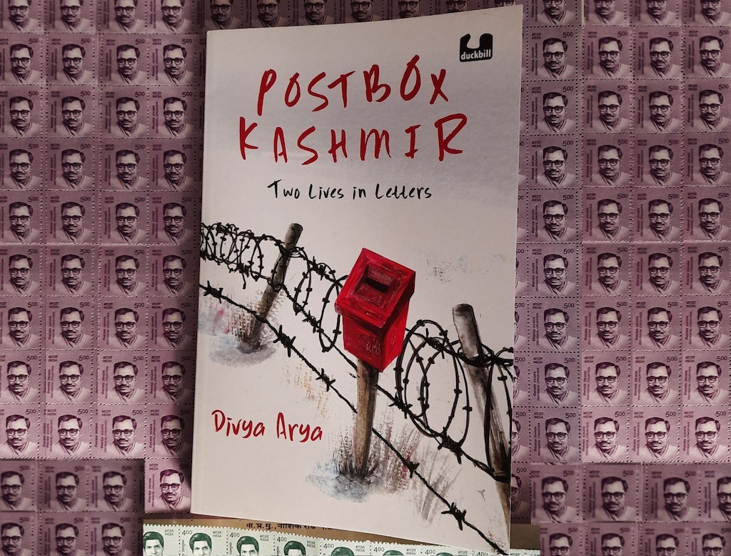 A book on a sheet of stamps. 
Text: Postbox Kashmir
Two Lives in Letters
Divya Arya
Image: A small red mailbox against a coiled barbed wire fence.